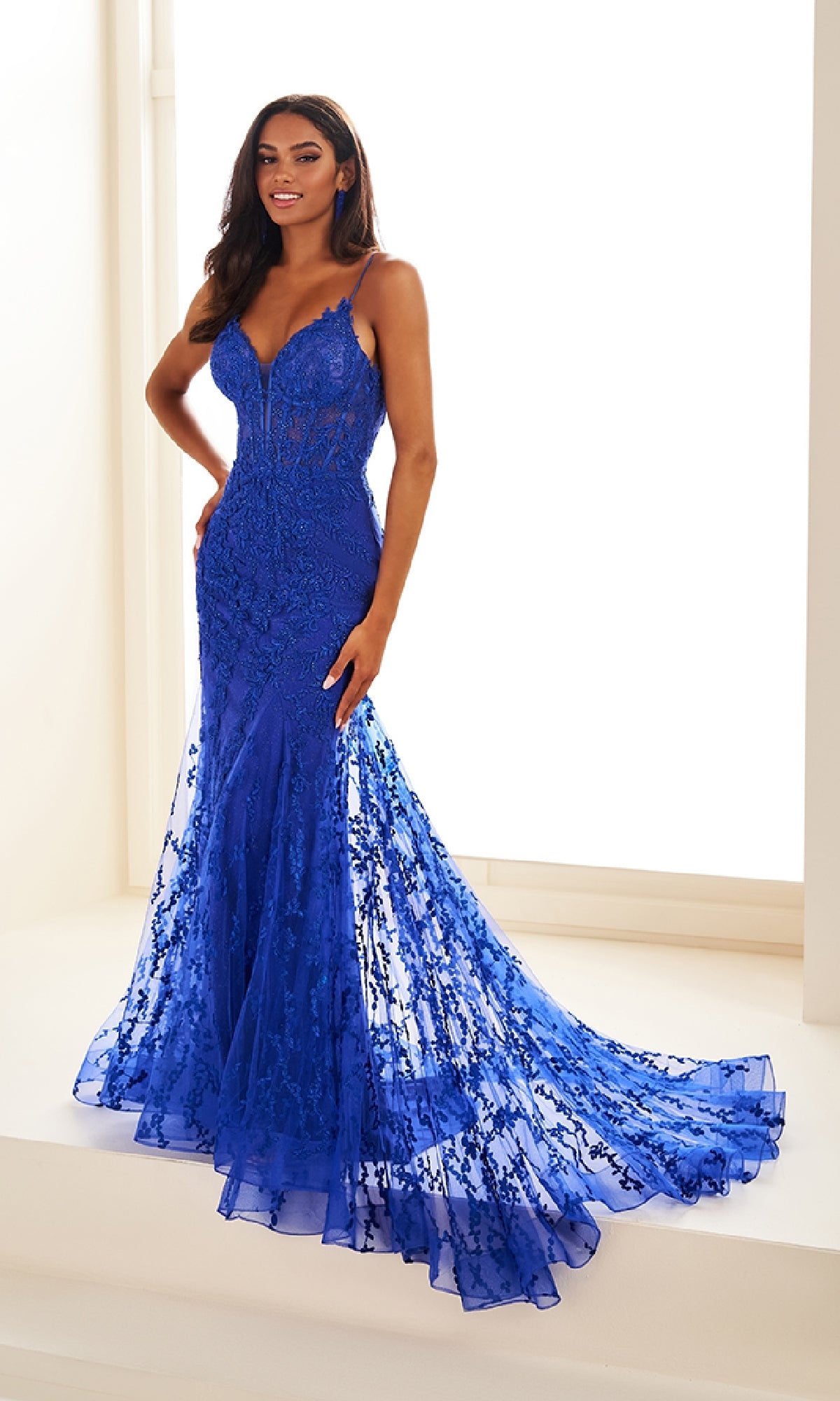 A woman stands indoors wearing the sleeveless, form-fitting Ellie Wilde Beaded-Tulle Long Prom Dress EW35010 in royal blue, featuring beaded embroidery, a mermaid silhouette, and a long train.